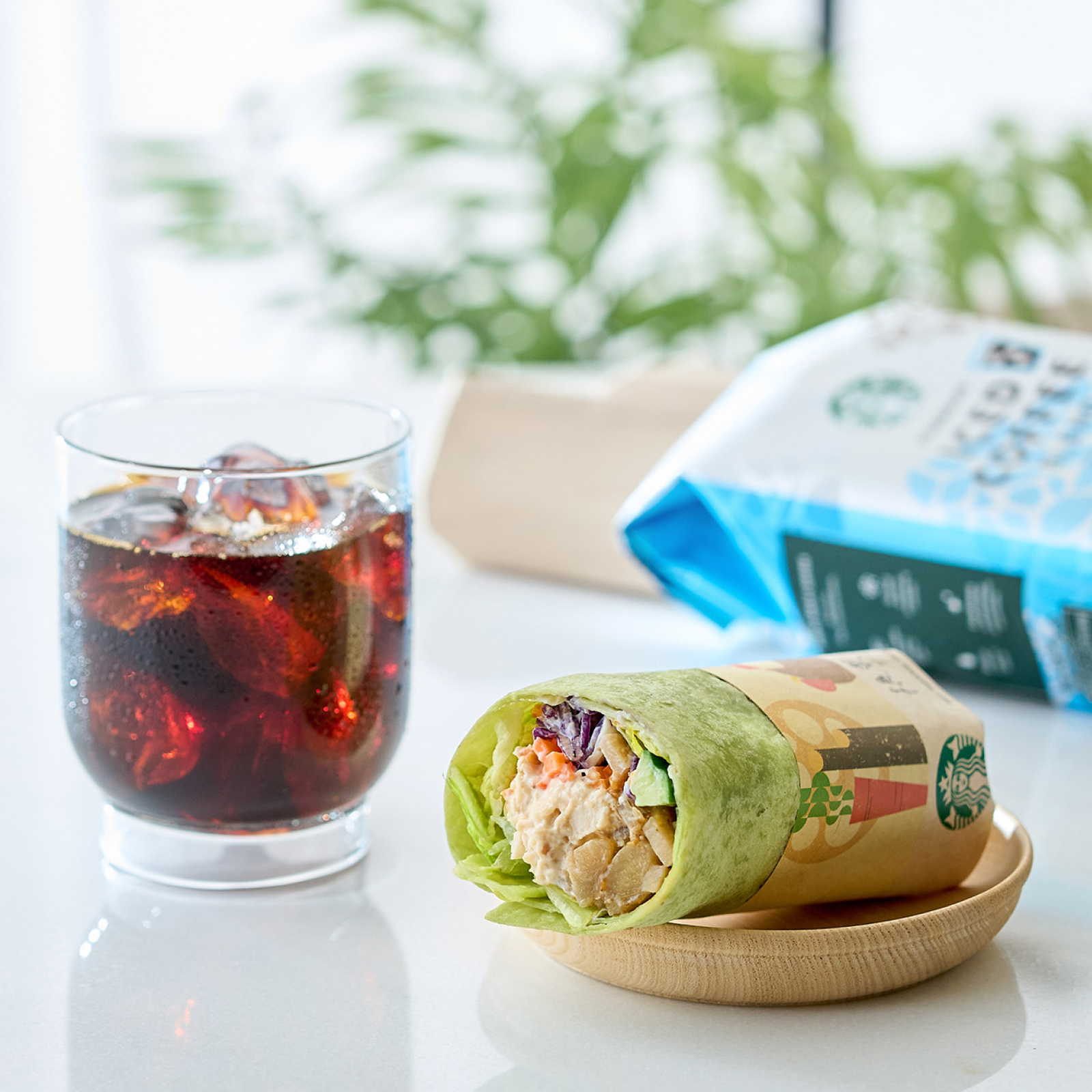 木目のお皿に置かれた根菜チキン サラダラップとスターバックス® アイスコーヒー ブレンドでいれたアイスコーヒー、スターバックス® アイスコーヒー ブレンドのパッケージ